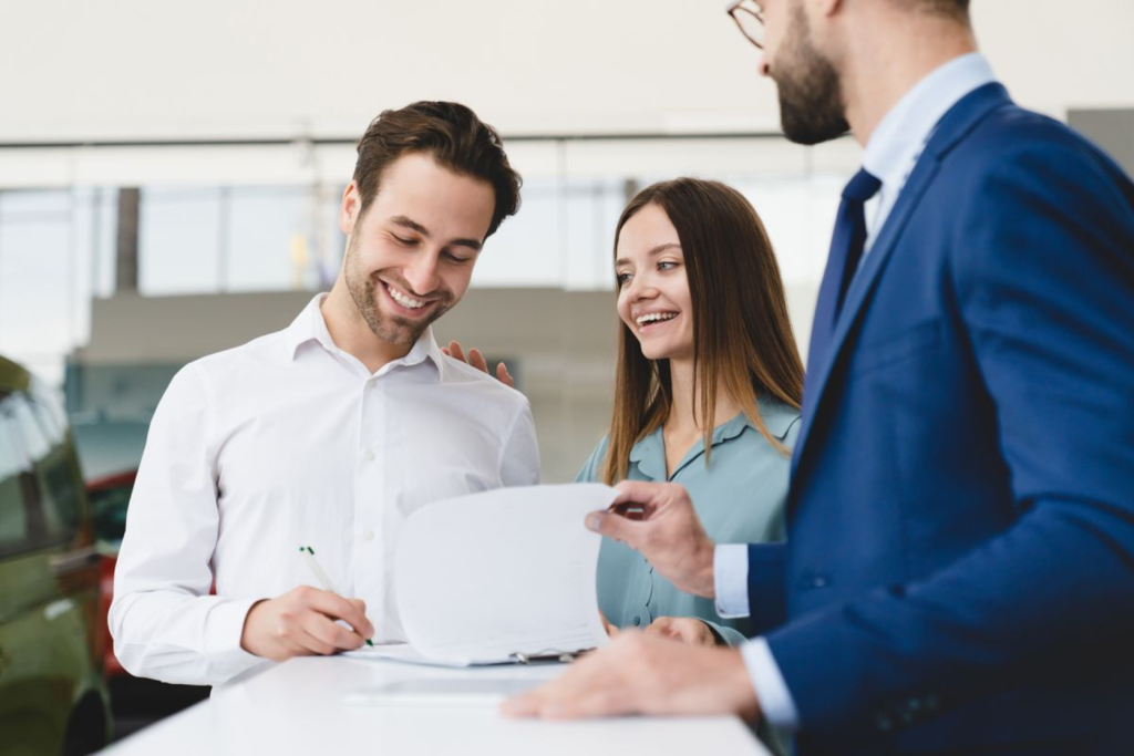 A couple smiling while signing paperwork at a car dealership with a sales representative.