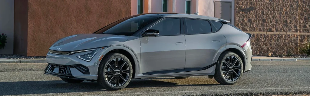 A silver Kia EV6 electric car parked on a street with a modern building in the background.